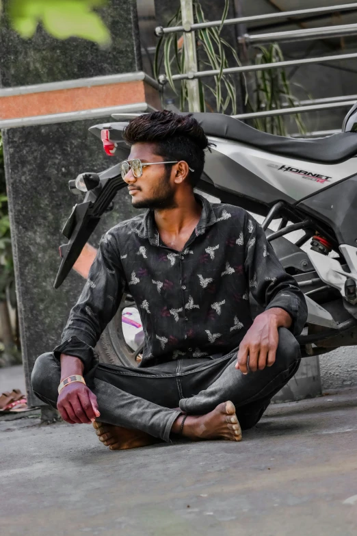 a man with glasses sitting on the street next to a motorcycle