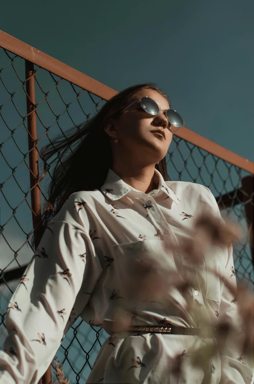 a woman with sunglasses, standing against a fence