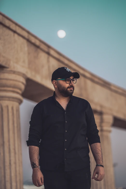 a bearded man with a black shirt and black hat walks through a cemetery in front of a large stone arch