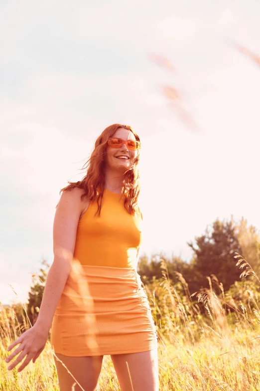 a smiling woman wearing an orange dress and sunglasses stands in the grass
