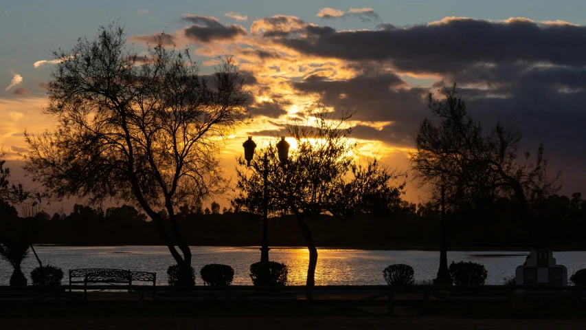 the sun is setting over a lake near a park bench