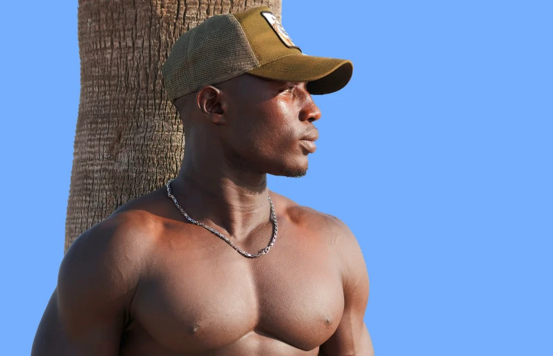 a shirtless male in a baseball cap stands against a tree