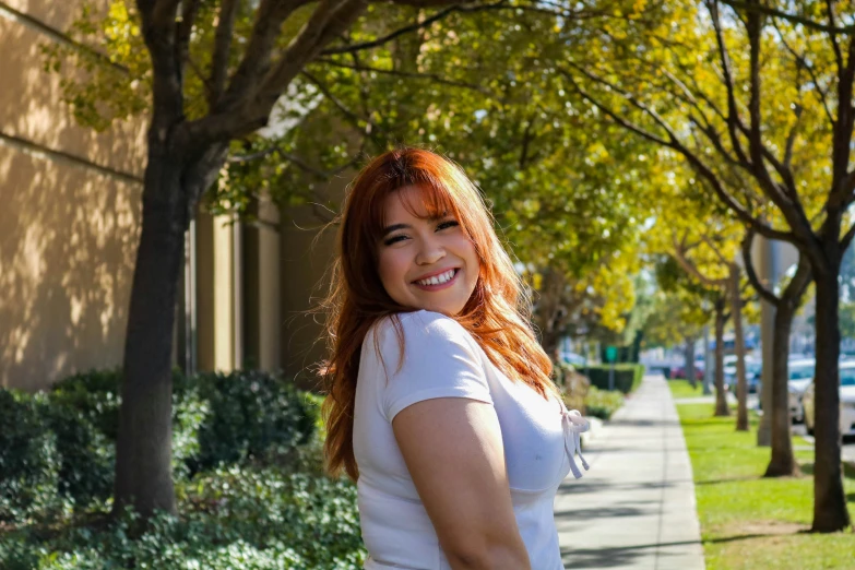 woman in a white top stands on a tree lined sidewalk