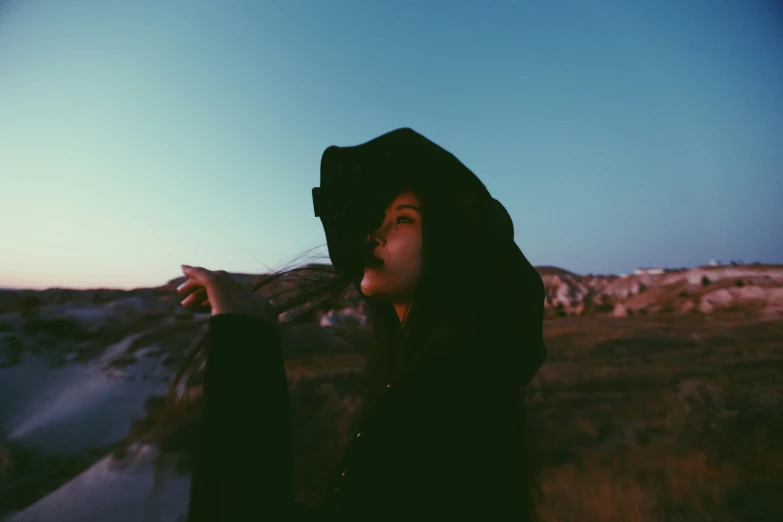 a woman in a dark hood and black jacket standing outside at dusk