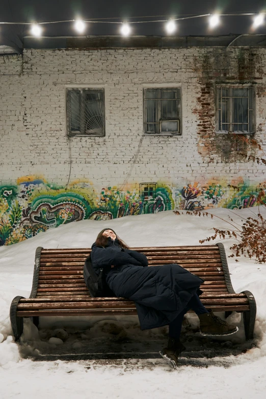 woman in long coat laying on a park bench covered with snow