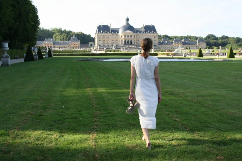 a woman wearing a white dress walking through the green grass of a palace