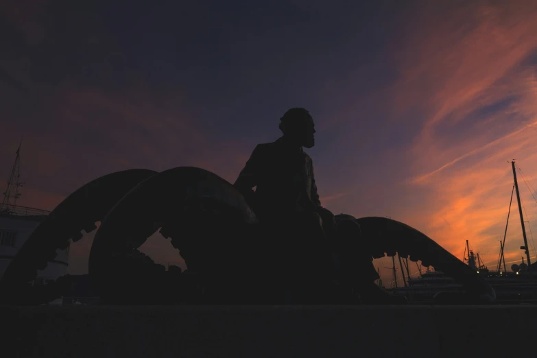 silhouetted figure sitting on the ground, watching the setting sun over boats