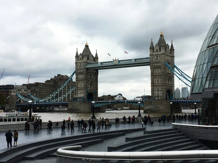 a group of people are walking next to the water in front of a bridge