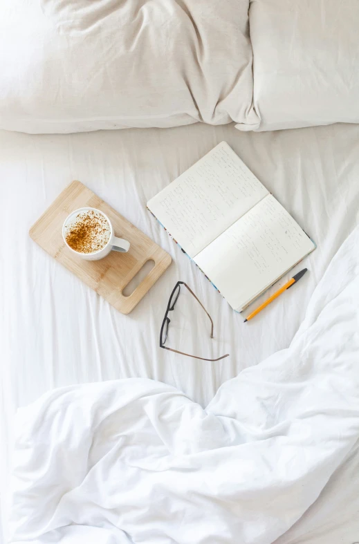 some glasses, an open notebook and a cup of coffee are set on a bed