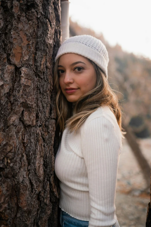 a young woman wearing a white hat leaning against a tree