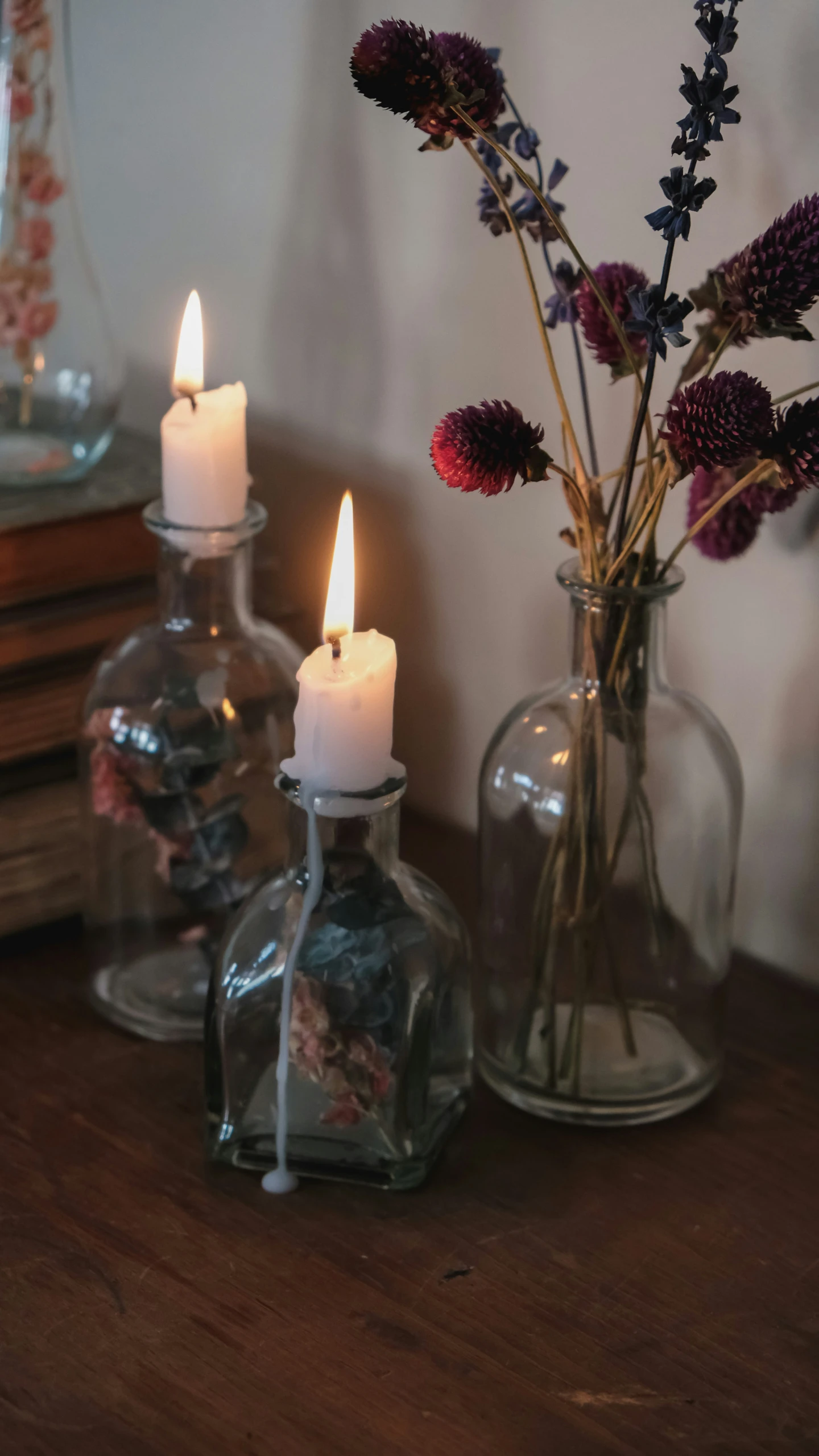 two glass bottles are on a wooden table, one with flowers in them