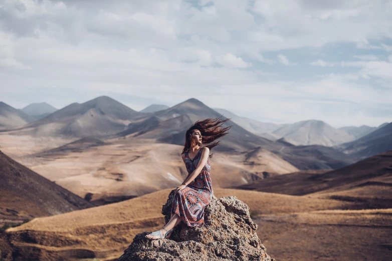 a woman with long hair sitting on a rock looking at the mountains