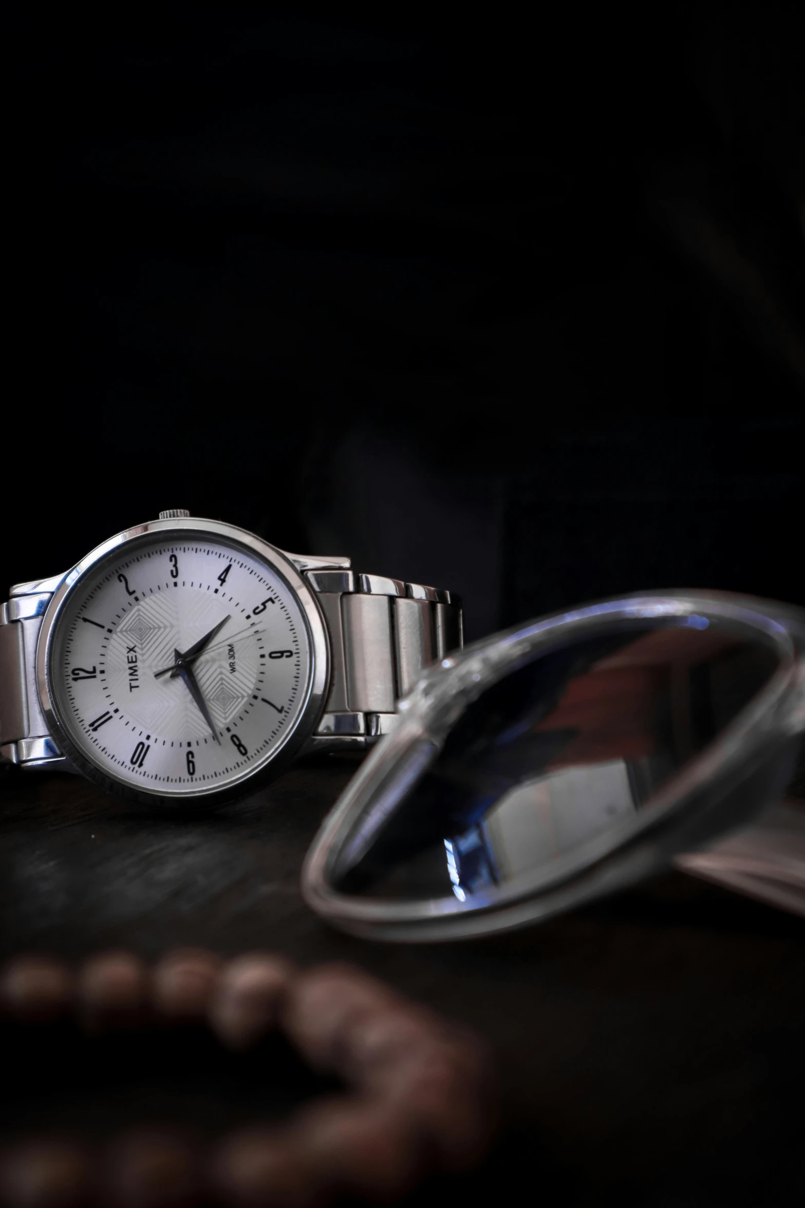 a round silver wrist watch sitting on a wooden table next to a pair of glasses