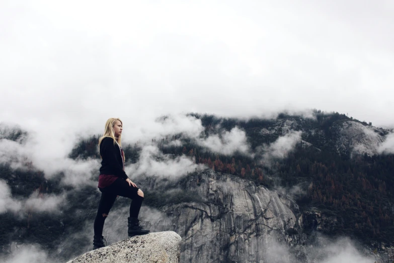 a woman stands on a cliff with clouds and mountains in the background