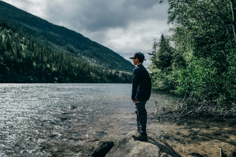 a man stands on a rock by the edge of a lake while looking off into a forest