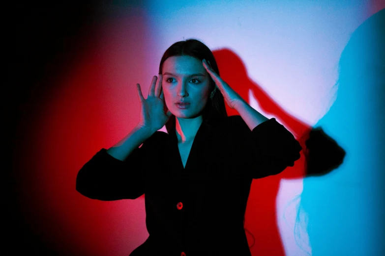 a girl with long hair, wearing black and standing in the shadows, is holding her hands on her ears