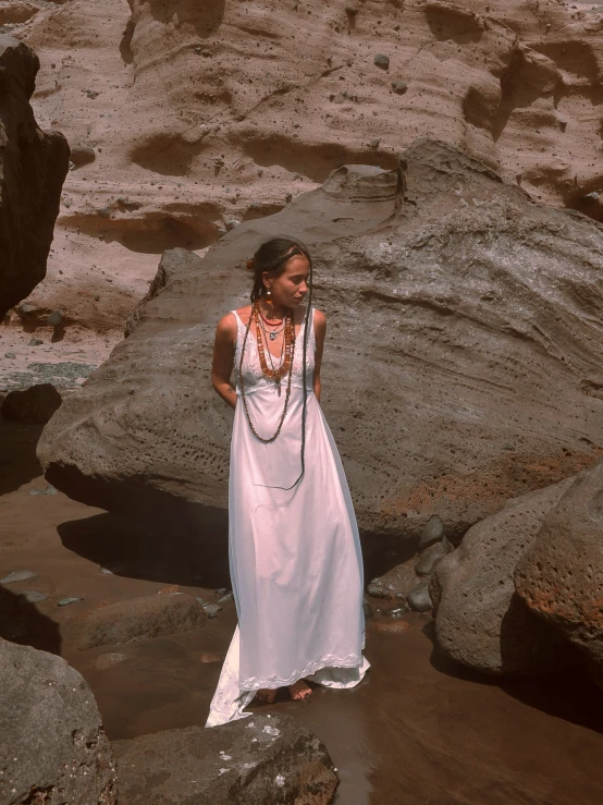 a woman wearing a long white dress standing next to rocks