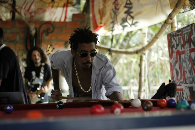 a man in sunglasses plays pool, while others look on