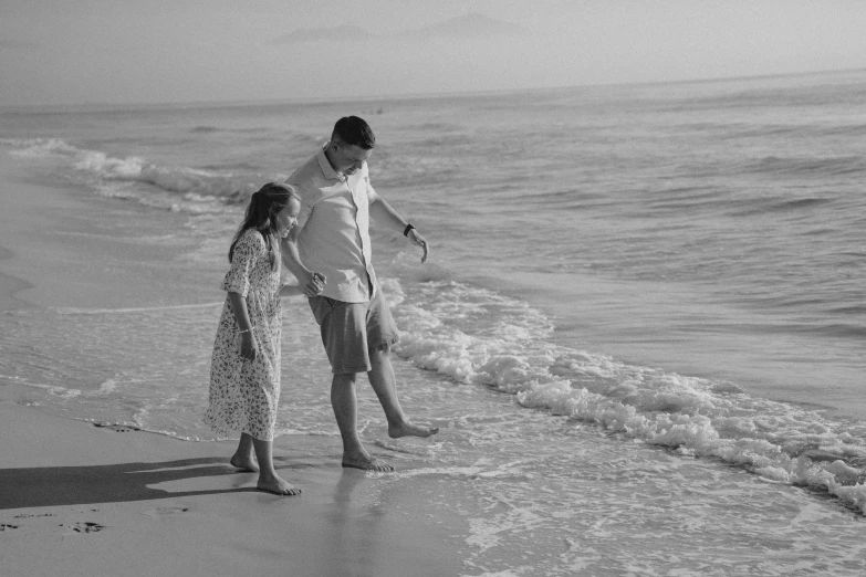 two s stand on the edge of the water as they walk along a beach with surf behind them