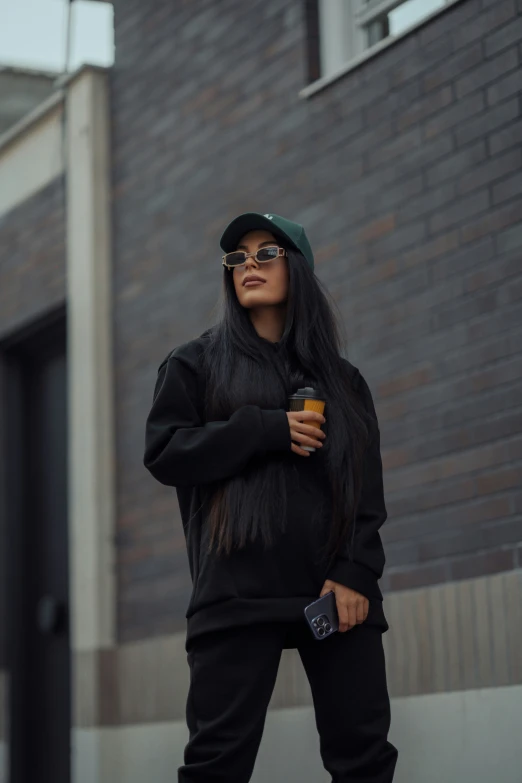 a woman in sunglasses and a beanie is standing on the sidewalk while holding a coffee cup