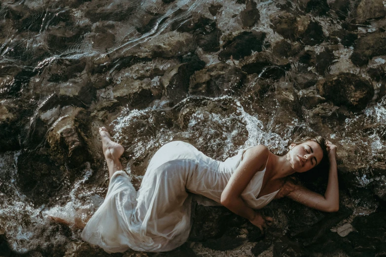 a woman lies on a rocky beach in white dress