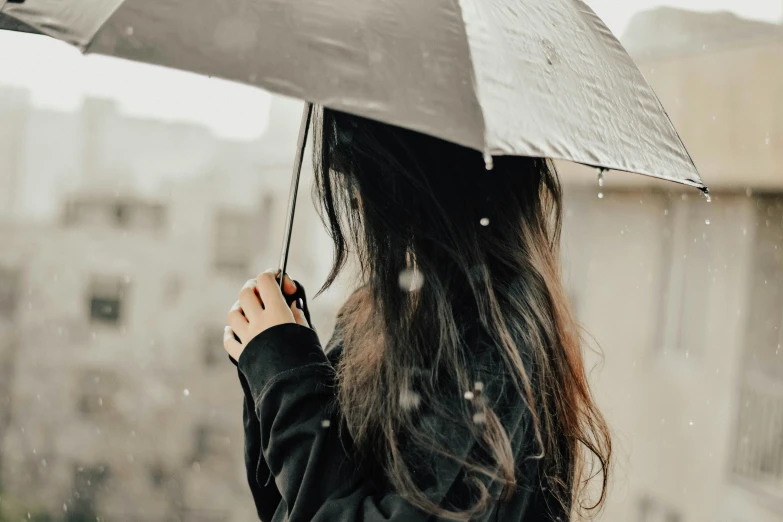 girl with long dark hair holding an umbrella looking over a rooftop at rain