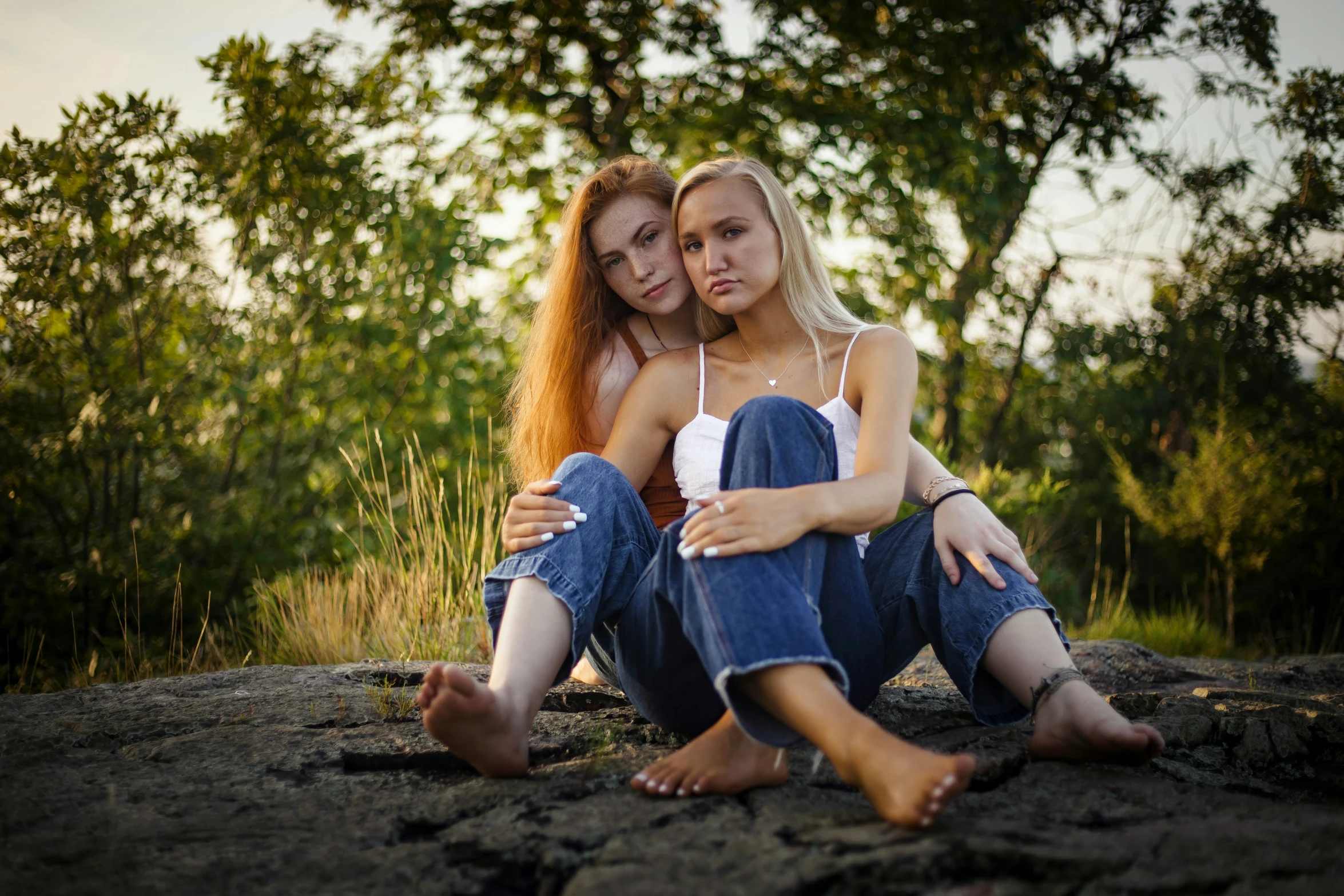 two women, each dressed in denims, sitting on rocks with trees in the background