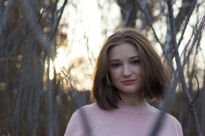 a young woman in a pink sweater stands next to tree nches