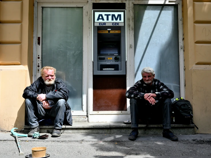 two men sitting outside a atm machine on the street