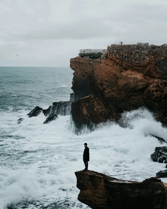 the man stands on the edge of a cliff next to the ocean as he looks at the crashing waves