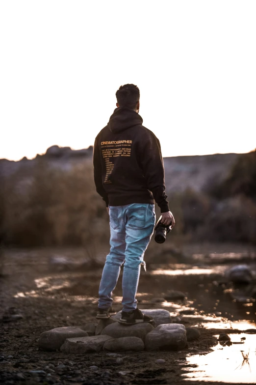 a man in a black jacket and blue jeans stands on rocks next to water