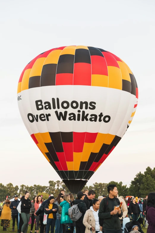 a huge balloon with a colorful stripe of red and yellow with the words balloons over waikato is flying in the air while people stand below it