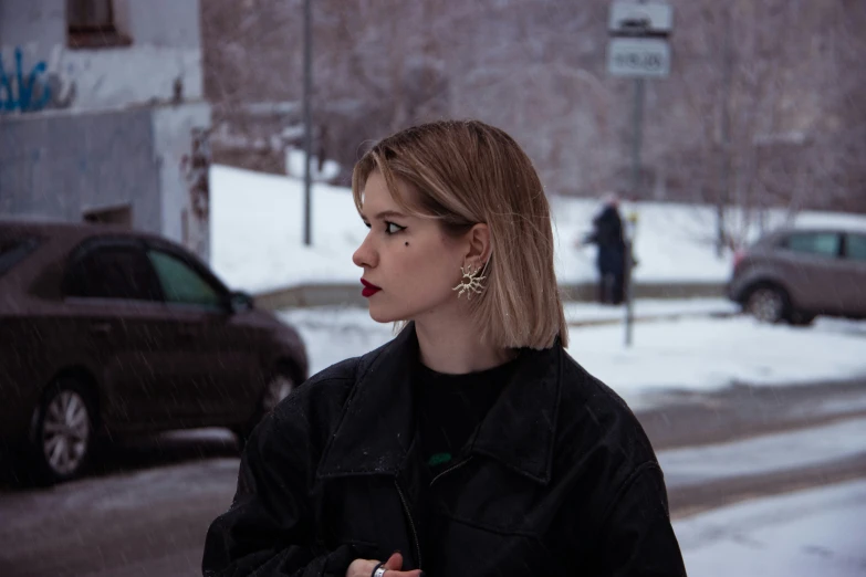 a woman in a black jacket and gold earring stand on the sidewalk in the snow