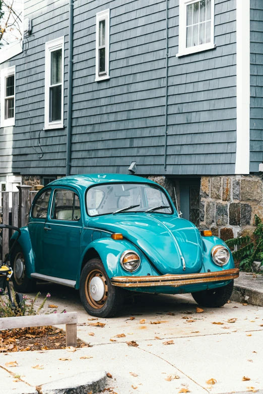 an old blue car sits in front of a grey house