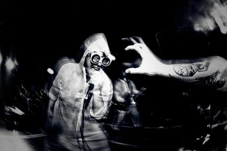 black and white pograph of a person wearing a gas mask with both hands on the wall and one hand behind his head