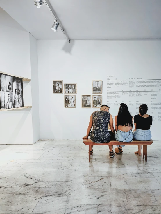 two women sit on a bench in a gallery, looking at framed pos