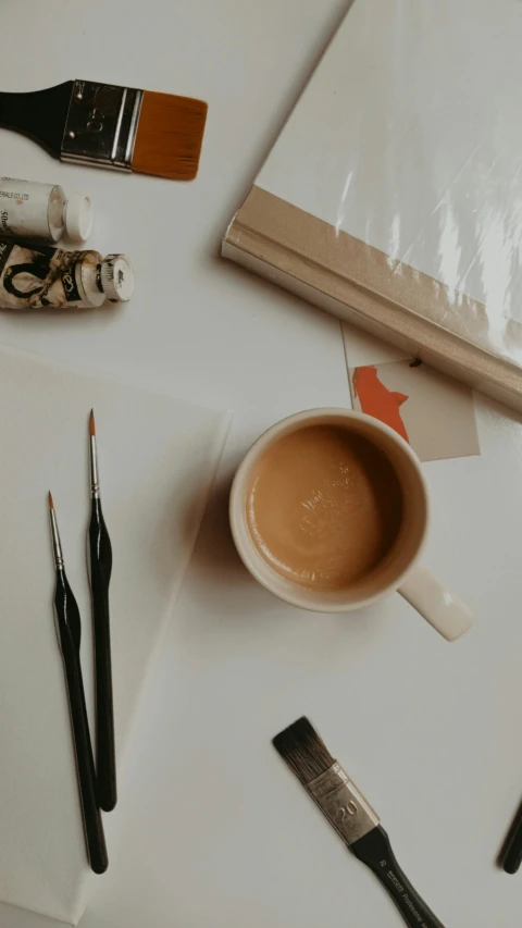 a coffee cup and three paint brushes sitting on top of a white table