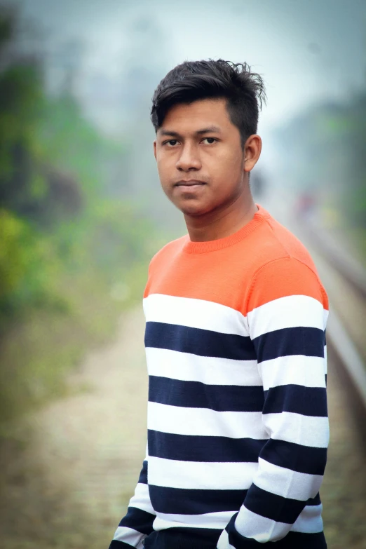 a young man with long dark hair stands in front of a train track and looks at the camera