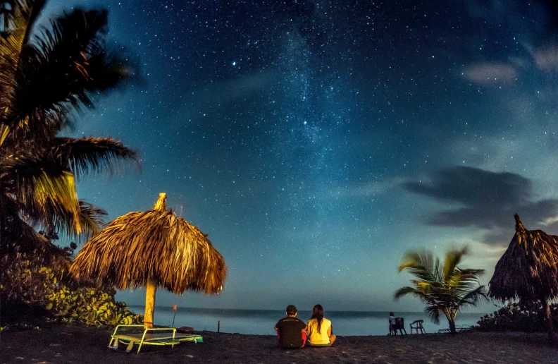 two people sitting on a beach under palm trees, looking at the stars and the night sky