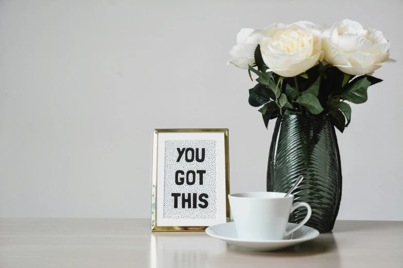 coffee mug, a white rose, and a framed sign sitting next to a vase with a few roses