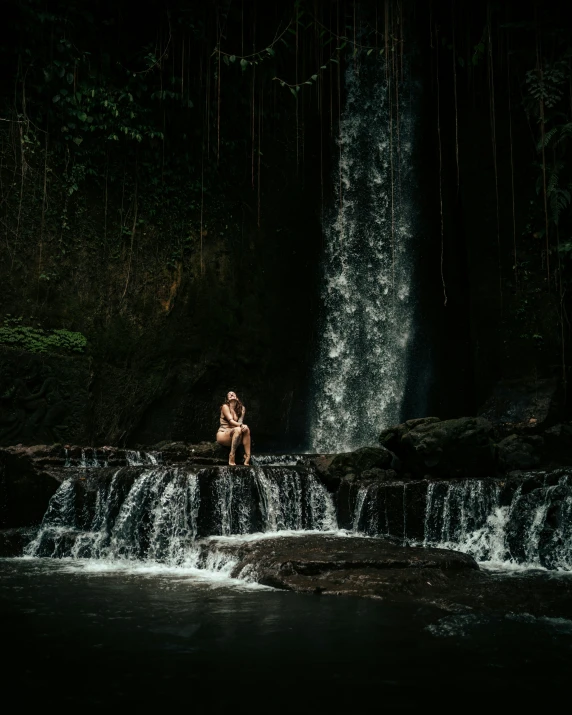 man kneeling on a rock in front of waterfall with water running down it