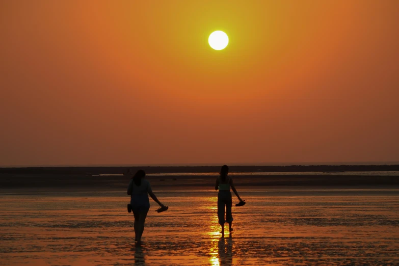 two people walk across a beach while the sun sets