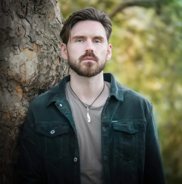 the man with a beard poses next to a tree