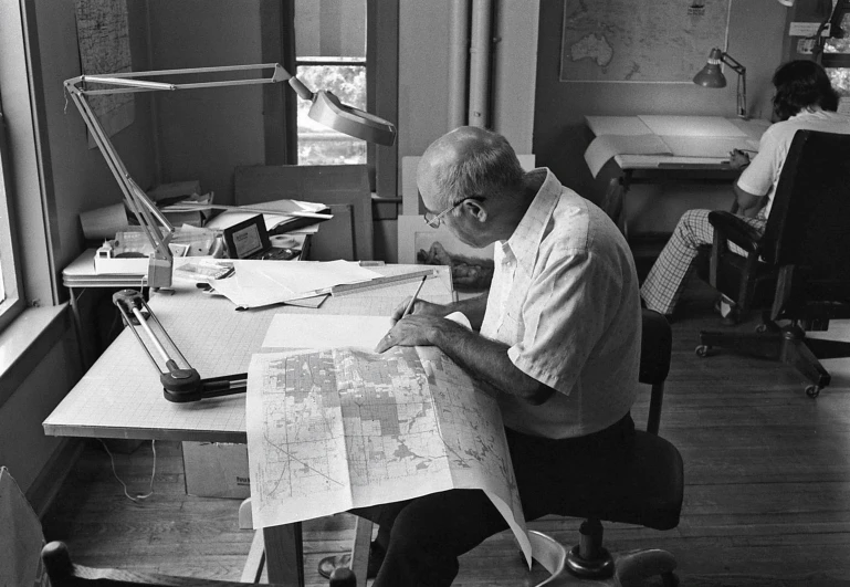 man sitting at desk looking over maps of land on top of him