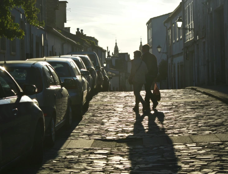 two people walk down a city street with parked cars