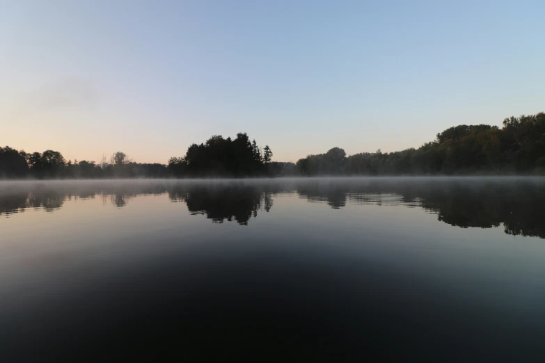 a lake surrounded by trees with fog rolling in the air