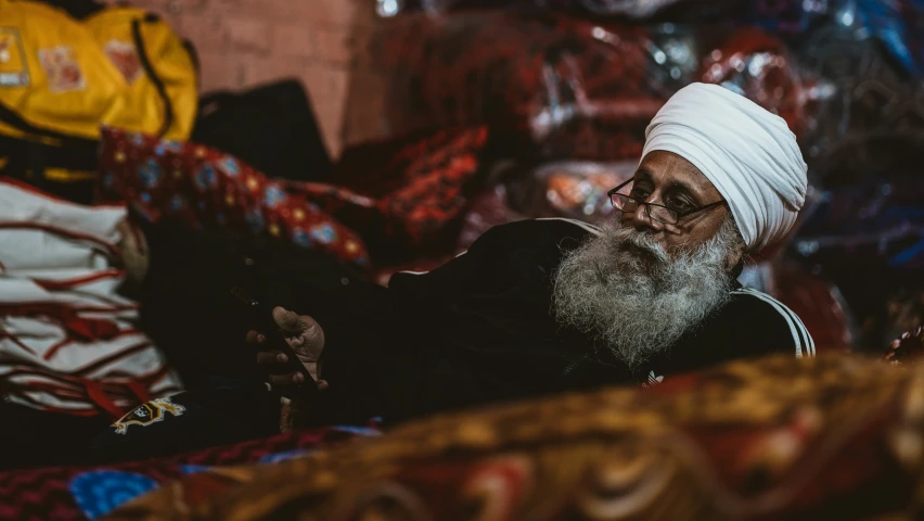 an old man with long white hair and a white turban is seated in a pile of cloths