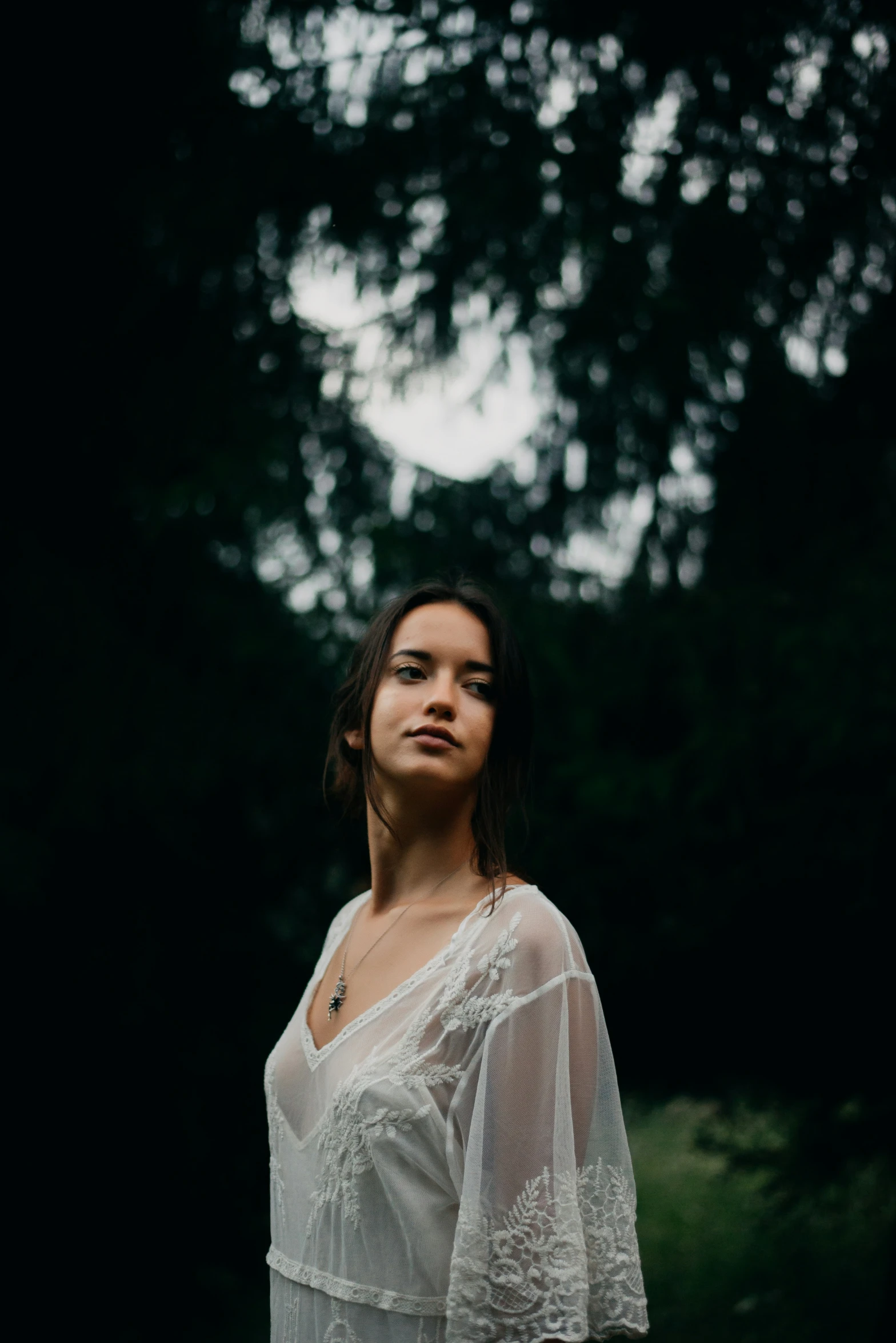 a woman wearing a white shirt and long black hair, standing in front of trees