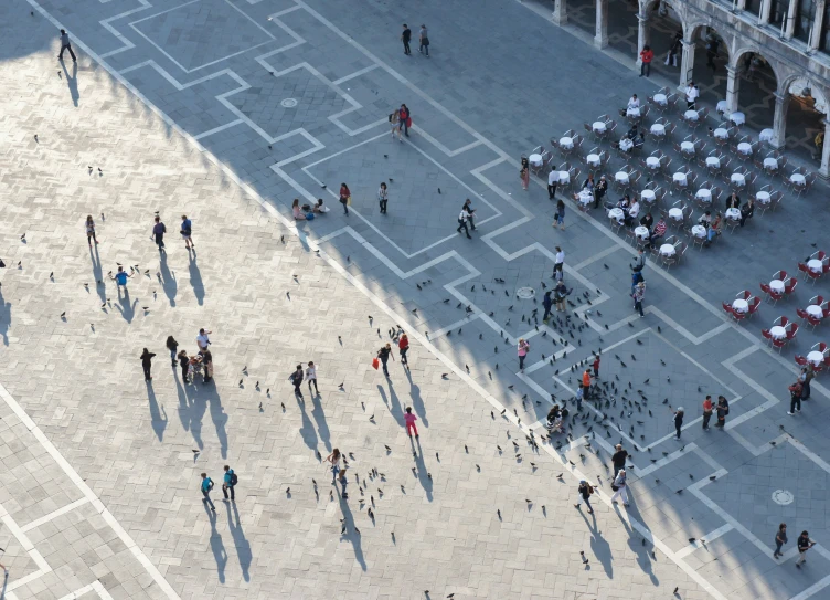 an aerial view of a group of people standing in a courtyard, looking down on tables with umbrellas open