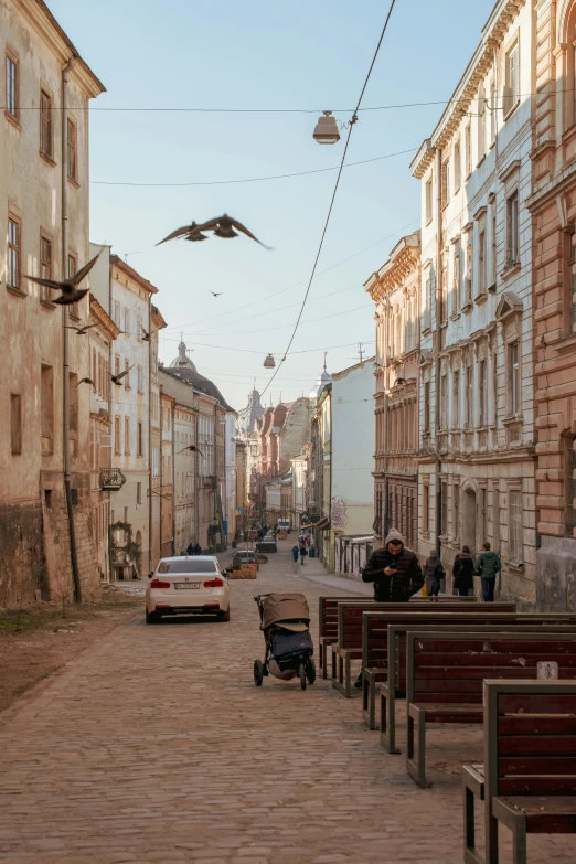 a street lined with old buildings with several empty park benches on one side, and one car on the other side with people walking on either side